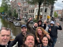 Group of smiling people posing near a canal with boats and historic buildings.