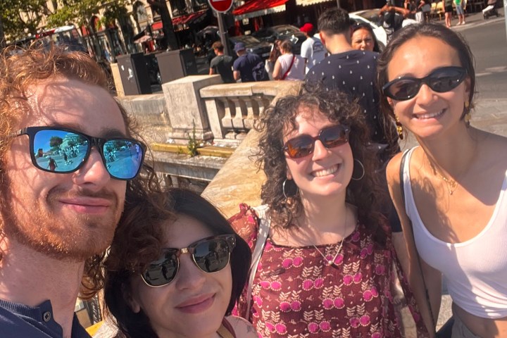 Four people smiling and posing for a selfie on a sunny street.