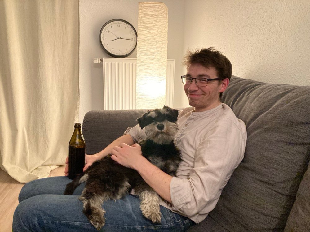 Person sitting on a couch with a small dog on their lap, holding a bottle, with a clock in the background.