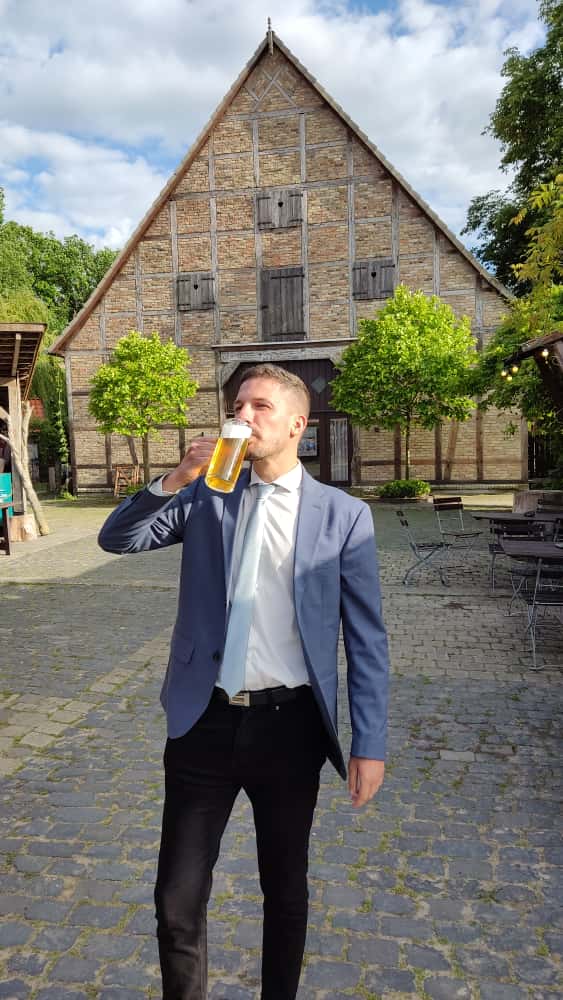 Man in suit drinking beer outside a rustic barn on a sunny day.