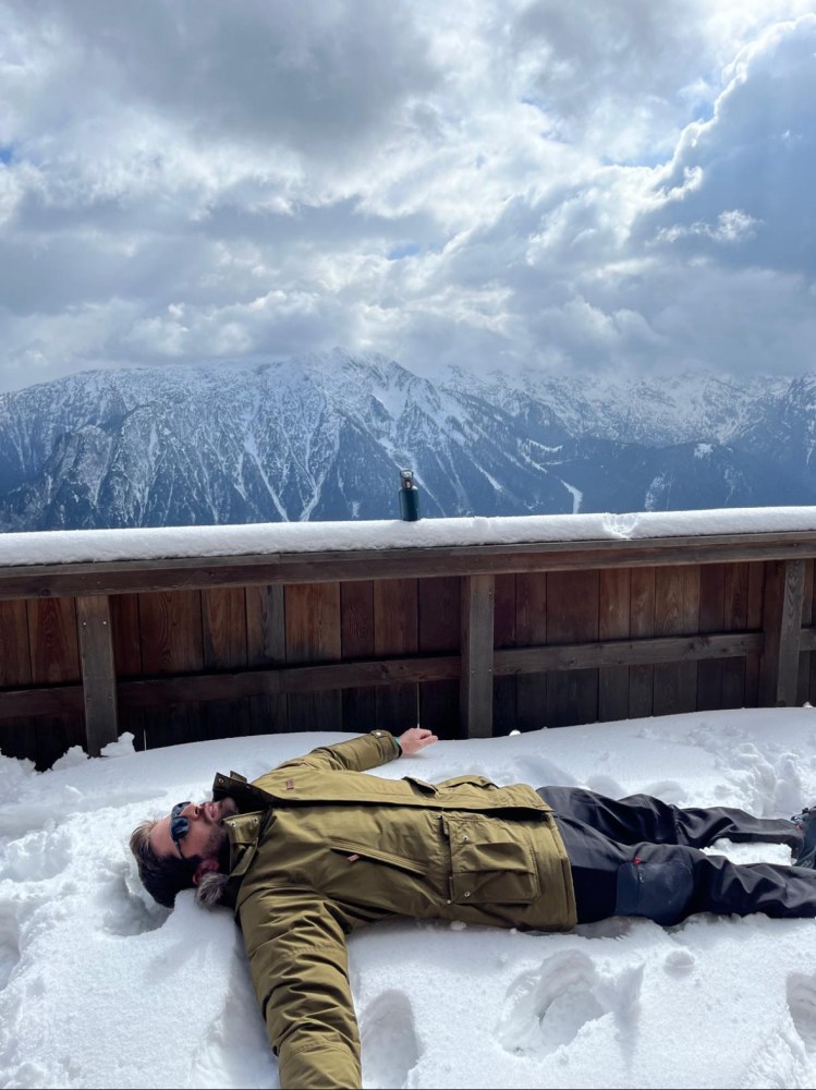 Person lying on snow, wearing a green jacket, with mountains and cloudy sky in the background.