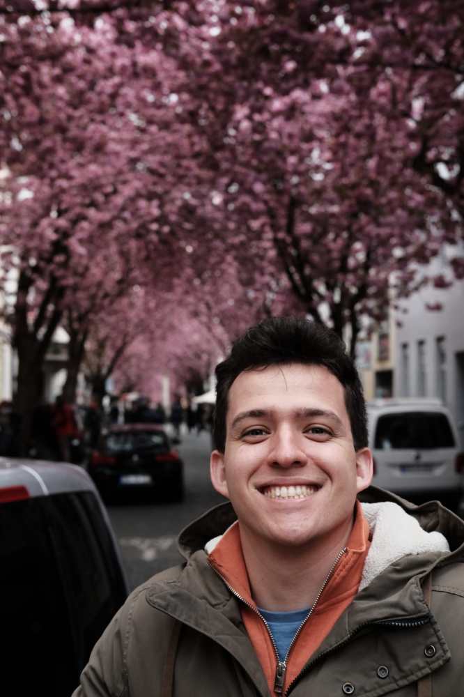 Smiling person in jacket with pink blossom trees lining a street.