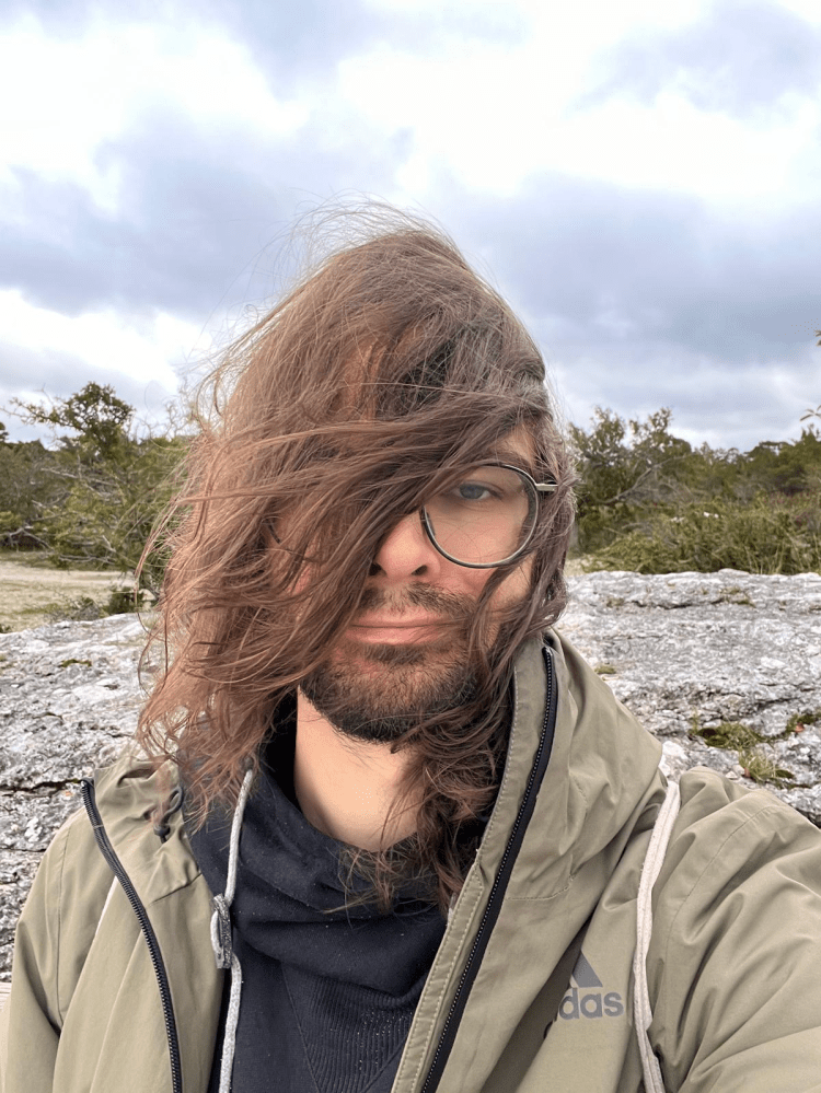 Person with windblown hair in glasses and jacket outdoors on a cloudy day.