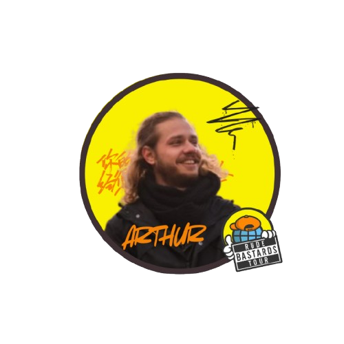 Person with long hair and beard against yellow background, black coat, text 'Arthurz', badge 'Rude Bastards Tour'.