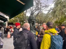 People gathered around a green fountain with trees and overcast sky.