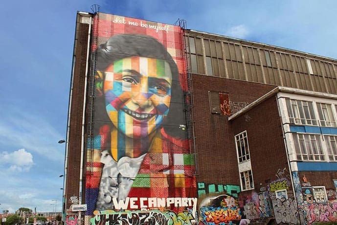 Colorful mural of a girl's face on a brick building, with graffiti at the bottom.