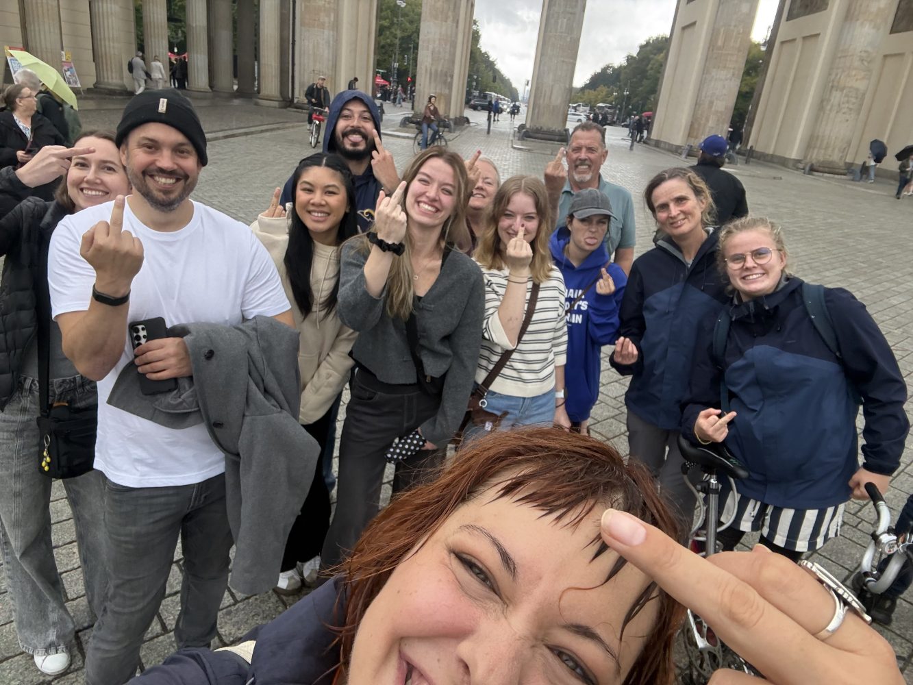 Group of people smiling for a photo outdoors, one person in front making a gesture.